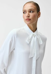 Biała bluzka z kołnierzykiem wiązanym na kokardę, długimi rękawami, gładką fakturą i zapinana na guziki z przodu. Prosty fason z delikatnymi akcentami.