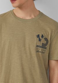 Olijfgroene katoenen T-shirt met een ronde hals en korte mouwen. Heeft een marineblauwe grafische print van palmbomen en tekst op de linkerborst.