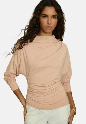 Light pink long-sleeve top with draped design and gathered texture at the waist, made of soft fabric. Worn with white pants.