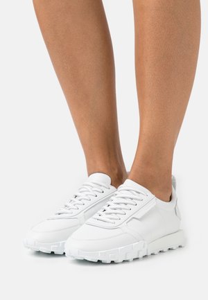 White athletic shoes made of smooth synthetic material, featuring a rounded toe, laces, and a textured rubber sole with prominent treads.
