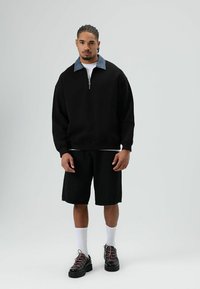 Zwart zip-up sweatshirt met denim kraag, gecombineerd met zwarte shorts. Model draagt zwarte schoenen met roze veters en witte sokken.