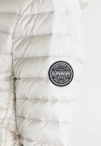 Lehká, nadýchaná béžová bunda se prošívaným designem a kulatou černou nášivkou s logem "SUPERDRY" na levém rukávu.