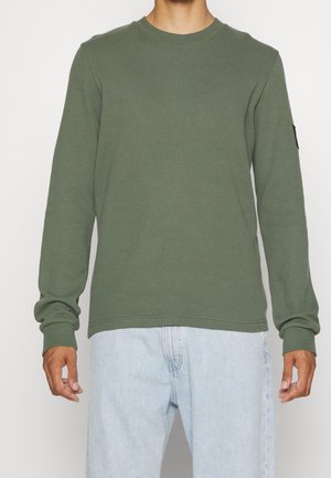 Homme portant une chemise en tricot gaufré à manches longues vert olive et un jean bleu clair, debout devant un fond blanc uni.