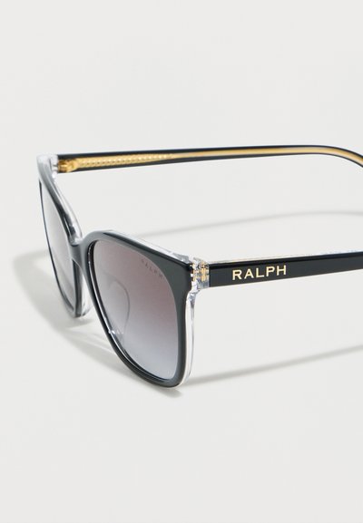 Γυαλιά ηλίου με μαύρη και διαφανή σκελετό, φακούς με διαβάθμιση χρώματος και χρυσή επωνυμία "RALPH" στα μαύρα πλαϊνά μέρη, φωτογραφημένα σε λευκή επιφάνεια.