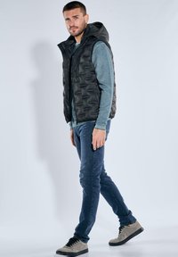 Schwarze Steppweste mit Kapuze über einem grauen Langarmshirt, kombiniert mit blauen Jeans und grauen Sneakers. Die Weste hat eine gesteppte Textur.