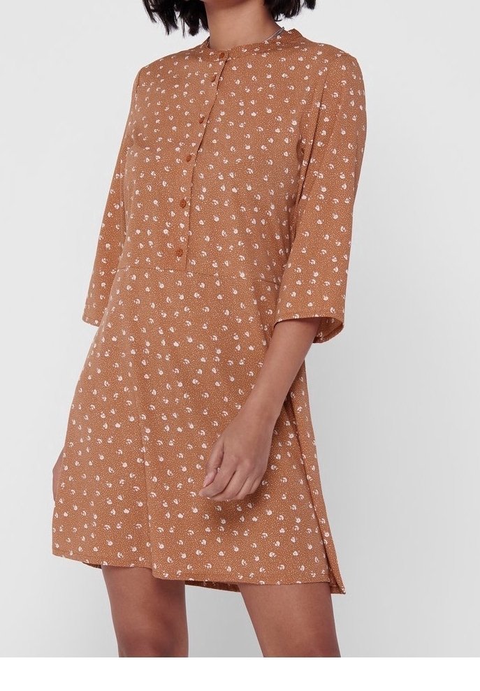 Femme portant une robe marron longueur genou avec imprimé floral rose, manches trois-quarts, et patte de boutonnage frontale sur un fond uni.