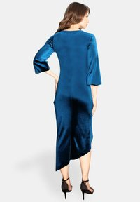 Abito in velluto blu con maniche a tre quarti, caratterizzato da un orlo asimmetrico e una texture liscia. Indossato con tacchi neri a strappi.