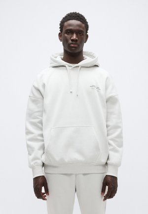 Gaiši pelēks pārizmēra džemperis no mīksta auduma, ar ķengura kabatu, regulējamu auklu un logotipu uz krūtīm.