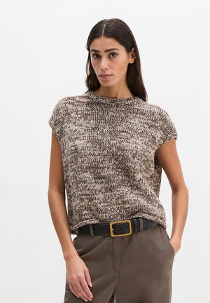 Frau mit langen dunklen Haaren, die ein braun-weißes, gestricktes ärmelloses Oberteil und braune Hosen mit einem schwarzen Gürtel mit goldener Schnalle trägt.