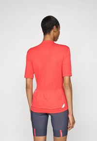 Rød idrettsskjorte med korte ermer og samlet midje, med en subtil tekstur. Kombinert med marineblå shorts med røde detaljer.