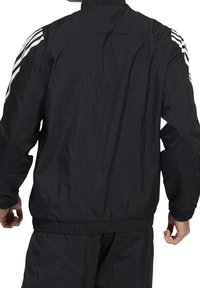 Svart lättviktsjacka med ståkrage, elastiska ärmar och tre vita ränder på varje axel. Skapad av ett strukturerat, vindtåligt material.