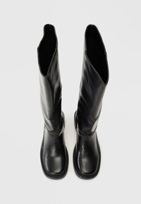 Bottes en cuir noir hauteur genou avec une texture lisse et une forme minimaliste. Détails cousus le long de la semelle et ouvertures extensibles.