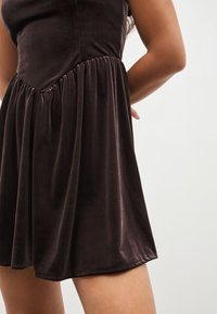 Temno rjava žametna obleka z oprijetim zgornjim delom, nabranim pasom in razširjeno krilom. Gladek tekstura z rahlim sijajem.