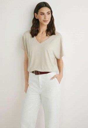 Kobieta z ciemnymi włosami do ramion, ubrana w beżowy top z krótkim rękawem i dekoltem w serek, włożony w białe spodnie z wysokim stanem i brązowy pas.