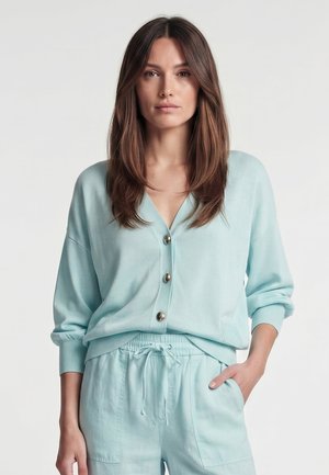 Frau mit langen braunen Haaren, die eine hellblaue, geknöpfte Strickjacke und passende Hosen mit Kordelzug trägt, steht mit einer Hand in der Tasche.