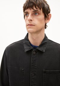 Schwarze Jeansjacke mit einem Kragen, vorderem Knopfverschluss und zwei Brusttaschen. Blauer gestrickter Pullover, der darunter sichtbar ist. Schlicher Hintergrund.