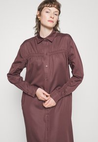 Vestido de botão bordeaux feito de tecido suave, com detalhes franzidos nos ombros e punhos, e um design de colarinho clássico.