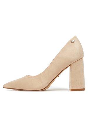 Zapato pump de ante beige con tacón alto, punta puntiaguda, tacón cuadrado y pequeño detalle de corazón dorado cerca del tobillo.