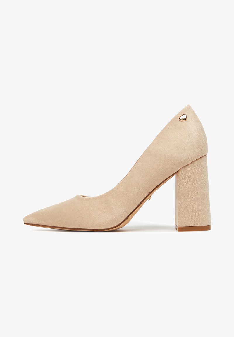 Zapato pump de ante beige con tacón alto, punta puntiaguda, tacón cuadrado y pequeño detalle de corazón dorado cerca del tobillo.