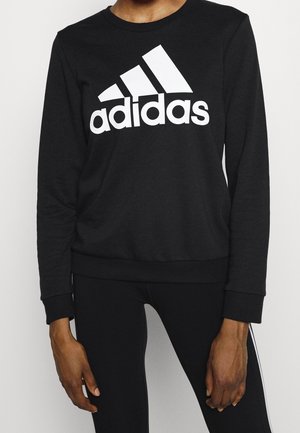 Persoon draagt een zwarte Adidas-trui met wit logo, gecombineerd met zwarte broek met witte strepen aan de zijkant.