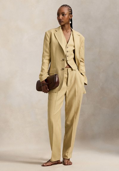 Ανοιχτό κίτρινο λινό κοστούμι που αποτελείται από σακάκι προσαρμοσμένο στο μέγεθος, γιλέκο και φαρδιά παντελόνια. Συνδυασμένο με καφέ τσάντα χειρός και φλατ σανδάλια.