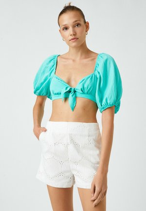 Türkises Crop-Top mit Puffärmeln und vorderer Bindeschleife, kombiniert mit weißen Shorts mit Muschelmuster und Lochstickerei.