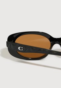 Mustad mustad prillid pruuni läätsega, millel on tekstuuriga käsi ja hõbedane "C" detail.