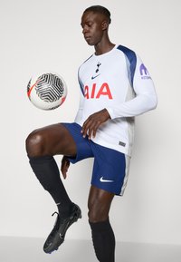 Hvid langærmet trøje med marineblå detaljer, med "AIA" i rødt, parret med teksturerede marineblå shorts og sorte fodboldstøvler, mens der dribles med en fodbold.
