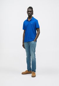 Kék pamut póló rövid ujjakkal, bordázott nyakkal, világoskék farmerrel és világosbarna hasítottbőr cipővel párosítva. Egyszerű dizájn, minták nélkül.