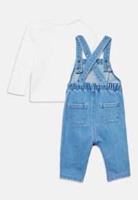 Λευκό πουκάμισο με μακριά μανίκια και στρογγυλή λαιμόκοψη, συνδυασμένο με μπλε τζιν σαλοπέτα που διαθέτει ρυθμιζόμενα λουριά και δύο πίσω τσέπες.