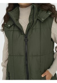 Woman wearing a sleeveless dark green padded vest over a cream cable-knit sweater, with one hand in the vest pocket.