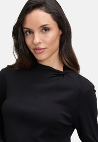 Top nero a maniche lunghe con collo alto e dettaglio a nodi sulla spalla. Tessuto liscio e elasticizzato che mette in risalto una silhouette aderente.