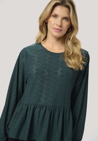 Türkisfarbener Langarm-Top aus weichem Material mit Peplum-Design, rautenförmigem Ausschnittmuster und rundem Halsausschnitt.