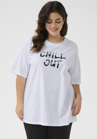 Camiseta de algodón blanca con cuello redondo y mangas cortas, que presenta un diseño impreso en negro que dice "CHILL OUT" en el frente.