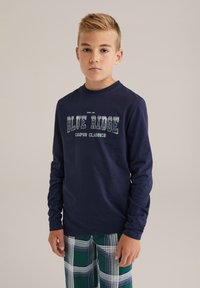 Jūras spēku garām piedurknēm krekls ar tekstu "BLUE RIDGE" un "CAMPUS CLASSICS" zem tā; kombinēts ar zaļām svītrainām biksēm. Mīksts kokvilnas materiāls.