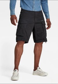 Calções cargo pretos com múltiplos bolsos e bainha ajustável, feitos de um tecido durável. Usados com uma camisa de ganga azul clara e ténis brancos.