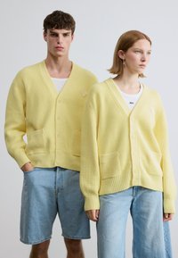 Κίτρινα ριγωτά ζακέτες με V-λαιμόκοψη, κουμπιά και εμπρόσθιες τσέπες, φορεμένα πάνω από λευκά μπλουζάκια. Σχέδιο ολοκληρώνουν ανοιχτόχρωμα τζιν σορτς.