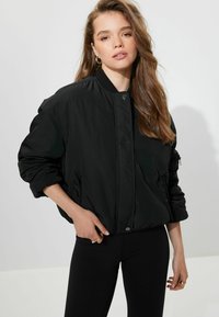 Veste bomber noire avec fermeture éclair sur le devant, poignets côtelés et poches latérales. Tissu doux et matelassé avec une finition légèrement brillante et coupe décontractée.