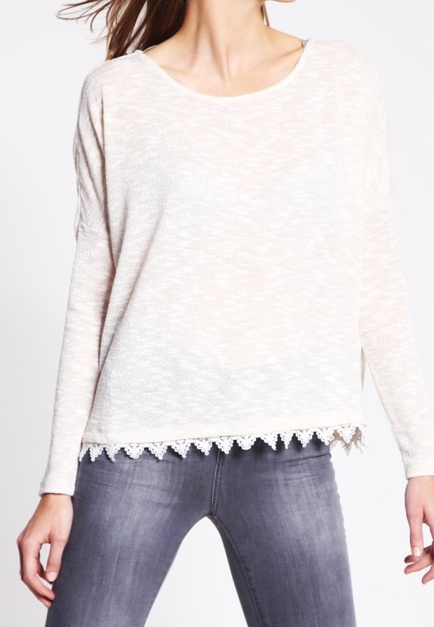 Femme portant un haut à manches longues beige clair avec une bordure en dentelle à l'ourlet et un jean gris moulant, debout les mains sur les hanches.