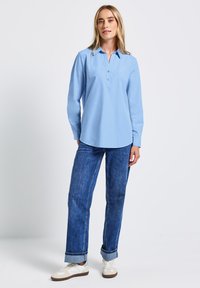 Camicia azzurro chiaro con maniche lunghe, colletto e orlo dritto, abbinata a jeans blu scuro e sneakers bianche.