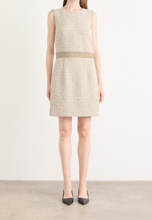 Vrouw draagt een mouwloze beige textuur jurk tot op de knie met een horizontale band in de taille en zwarte puntige hoge hakken.