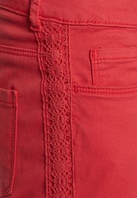 Pantaloni rossi con un bordo in pizzo laterale, caratterizzati da una parte frontale piatta e tasche posteriori, texture liscia e cuciture a contrasto lungo le cuciture.