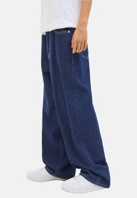 Personne portant un jean en denim bleu foncé surdimensionné et des baskets blanches, se tenant sur un fond blanc uni. La main repose sur le côté.