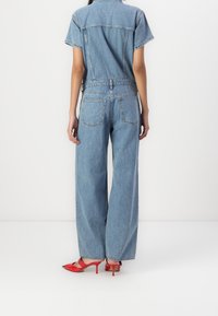 Mono de mezclilla de manga corta, lavado azul claro, corte relajado. Presenta dos bolsillos traseros, combinado con zapatos de tacón rojos.