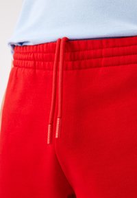Røde sweatpants med en strækbar talje, der har en snor med røde plastikspidser prægede med "Lacoste." Glat bomuldsstof.