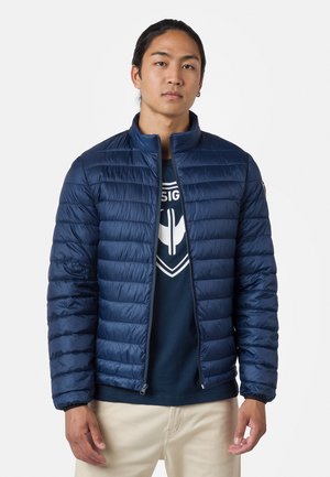 Marineblauwe gewatteerde jas met verticale quiltpatroon, staande kraag, ritssluiting en elastische manchetten. Draagt boven een donker T-shirt.