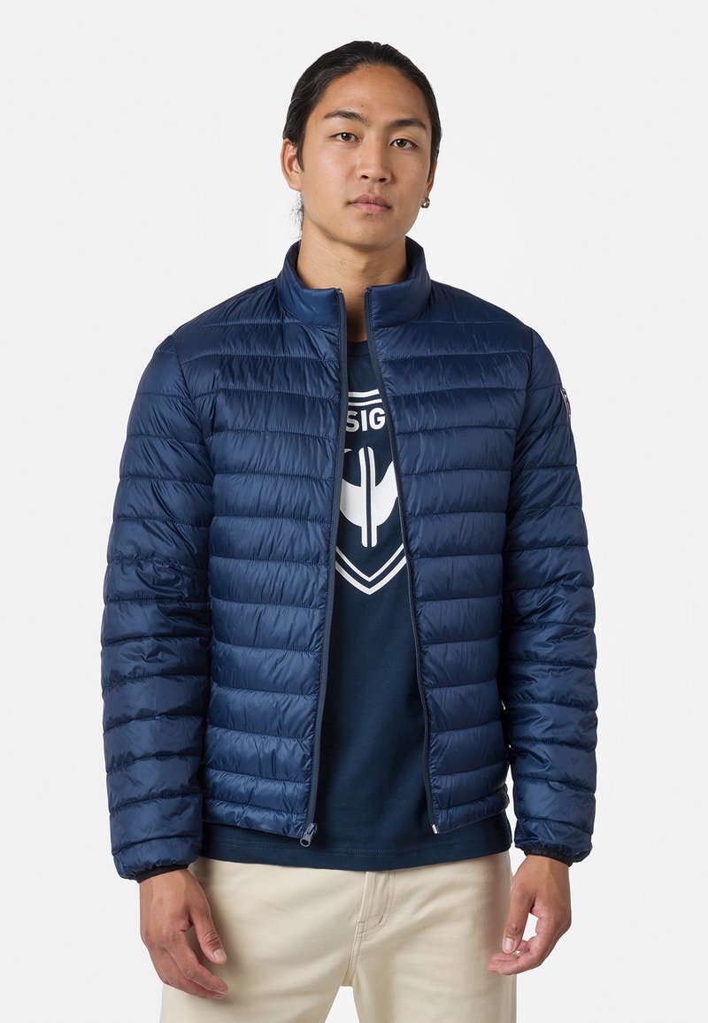 Marineblå puffjacka med vertikalt quiltat mönster, ståkrage, dragkedjestängning och elastiska ärmslut. Bärs över en mörk T-shirt.