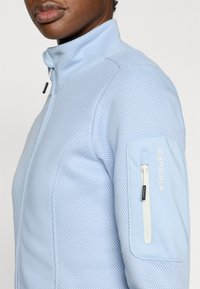 Αθλητικό τζάκετ ανοιχτού μπλε με υφή κυψελής. Διαθέτει ψηλό γιακά και φερμουάρ σε θήκη στο μανίκι. Ανθεκτικό ύφασμ α με διακριτικές λεπτομέρειες.
