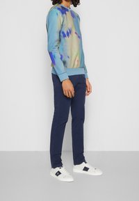 Felpa unisex multicolore in tie-dye in morbido tessuto con polsini a costine, abbinata a chinos blu navy e sneakers bianche con dettagli blu.