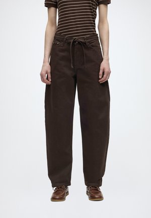 Άτομο που φοράει φαρδιά σκούρα καφέ παντελόνια με κορδόνι στη μέση, καφέ ριγέ κοντομάνικο πουκάμισο και καφέ δερμάτινα παπούτσια σε λευκό φόντο.